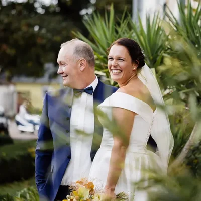 Hochzeitspaar steht zwischen Palmen, die Braut strahlt in natürlicher Hochzeitsatmosphäre